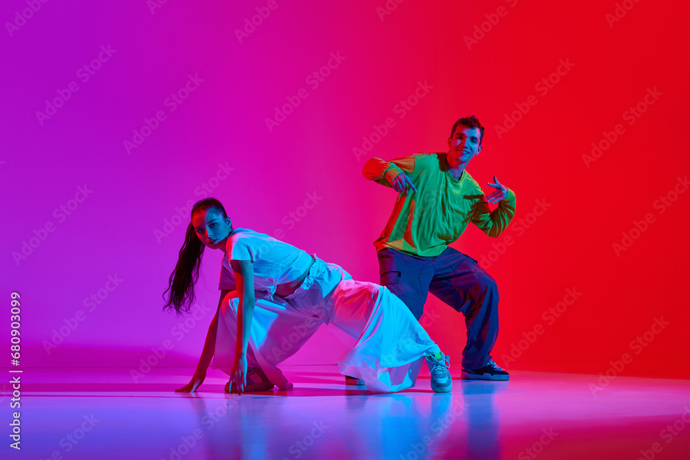Creative flow. Young man and woman in motion, dancing hip hop against pink red background in neon light. Concept of hobby, action, street style, contemporary dance, youth, fashion