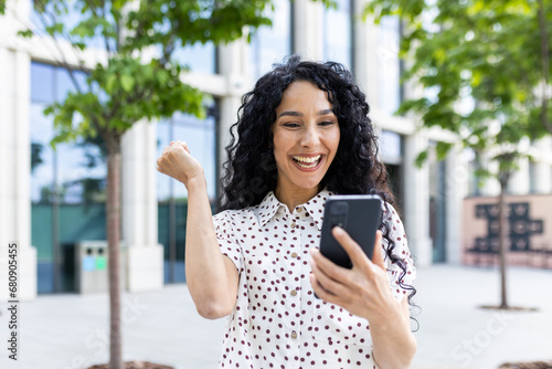 Young joyful woman winner received online notification on phone, Hispanic woman with curly hair celebrating success and triumph walking in city near office building outside photo
