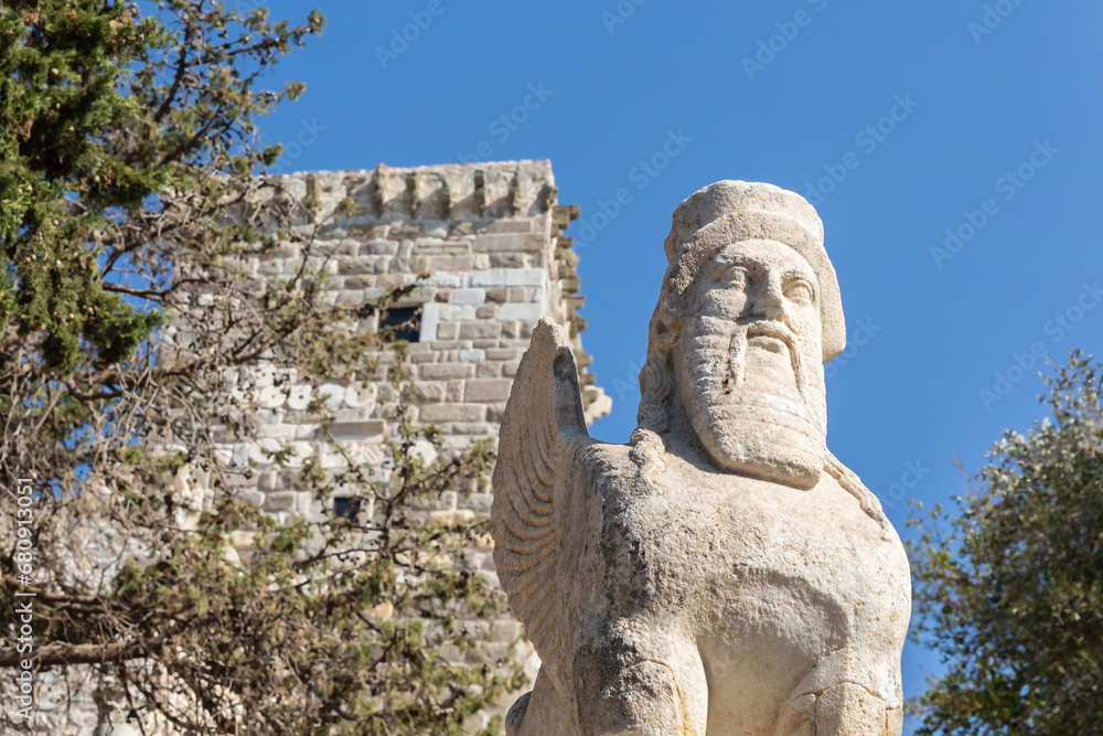 Statue of the ancient winged sphinx with the head of a bearded man and the medieval tower and blue sky at background. Bodrum (Mugla), Turkey (Turkiye)