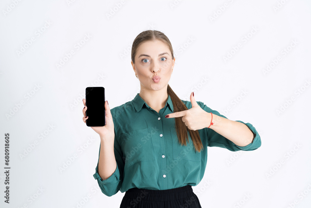 Attractive young woman is surprised and points her finger at an empty mobile phone display for a copy space for advertising isolated on a white studio background. A concept smartphone. application Attractive young woman is surprised and points her finger at an empty mobile phone display for a copy space for advertising isolated on a white studio background. A concept smartphone. application