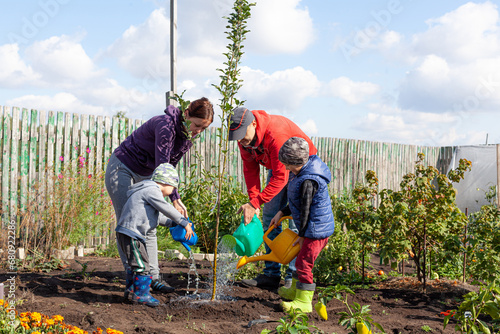 A family of four: mom, dad and two sons watering a young, freshly planted tree from a watering can on an autumn day