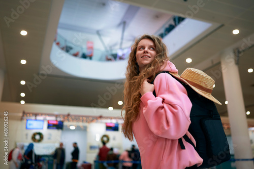 Joyous woman with backpack and hat anticipates holiday journey inside busy airport adorned for Christmas. Festive travel vibes, seasonal wanderlust.