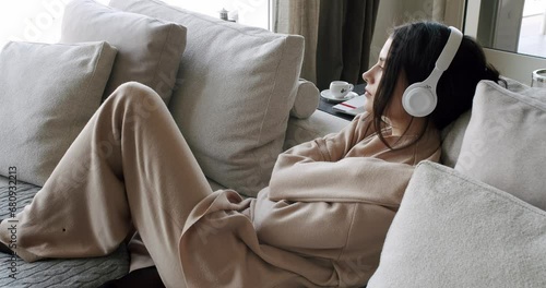 Sad young woman feeling loneliness, lying on couch in wireless headphones and listening relaxation music hugging yourself. Thoughtful female resting on sofa. Depression concept. Melancholy mood photo