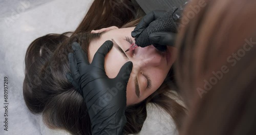 Beautician in black gloves making permanent makeup of eyebrows to young client woman in beauty salon. Cosmetologist female with professional tool doing microblading procedure, brow tattoo.  photo