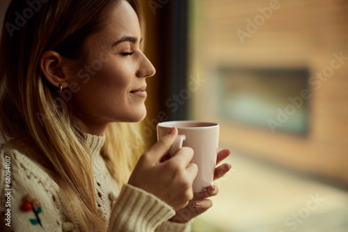 Side view of a smiling girl, keeping her eyes closed, standing near the window, holding a hot coffee.