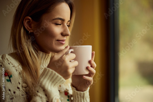 A smiling girl, keeping her eyes closed, holding a cup of tea, being indoors.