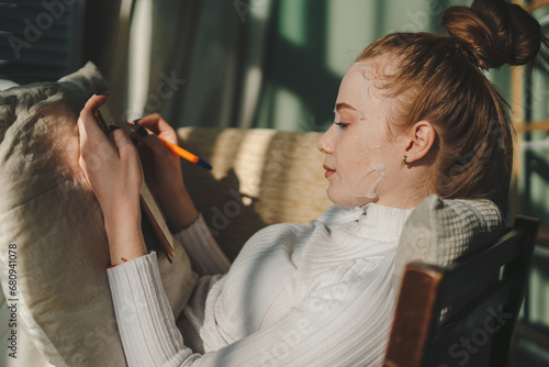 Side view of a beautiful young ginger woman early in morning, writing notes in notepad, sitting on comfortable sofa. Cozy domestic atmosphere.