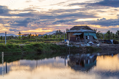 An old shack, located by a river in central Vietnam. The sun is setting through a cloudy sky.