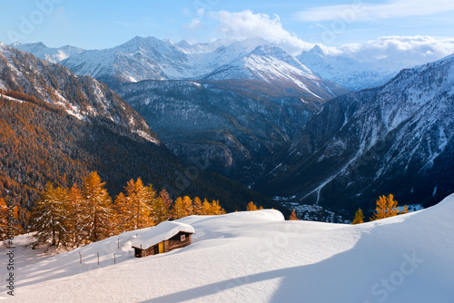 Small wooden cabin on snowy meadow on sunrise time. Courmayeur, northern Italy, Italian Alps. Snowy hills with orange larch forest and Monte Bianco mountains group. Landscape photography