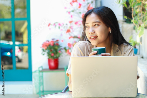 happy asian woman drink coffe while work in cafe with laptop sit in outdoors wearing casual clothes fashionable, female remote worker smiling look away
