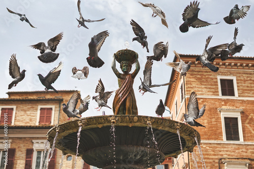 Osimo, Italy: Fontana della pupa. Famous ancient fountain located in Boccolino square. Several pigeons flying over a water fountain. Birds cool off in the summer. Pigeons bathing.