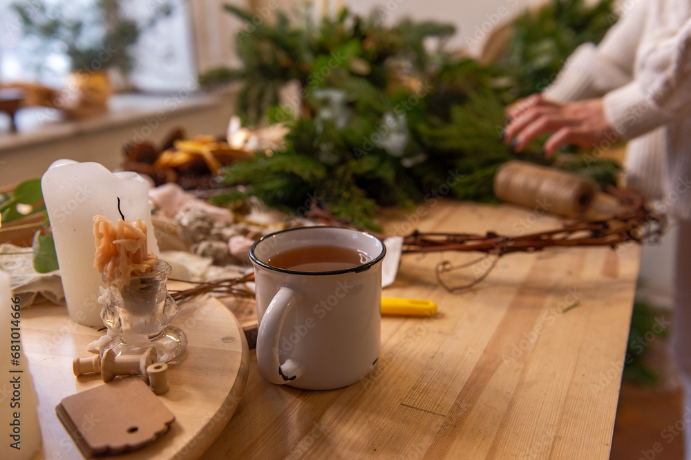 Side view still life, flat lay female hands holding white cup of tea on wooden table. Place for text, on background are tools, natural materials for making handmade Christmas wreath. Copy space