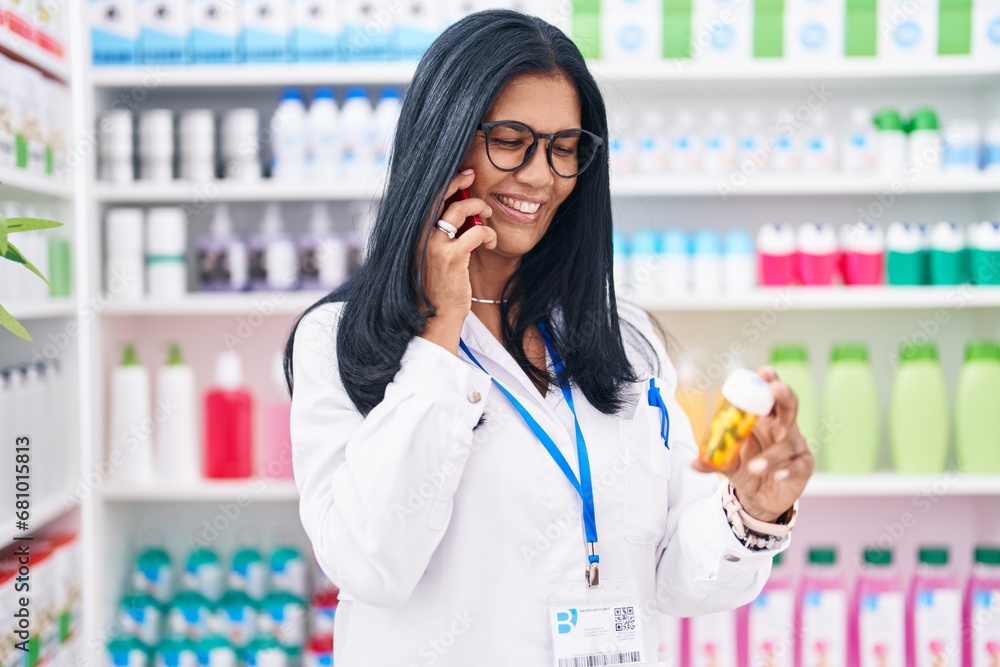 Middle age hispanic woman pharmacist holding pills bottle talking on smartphone at pharmacy