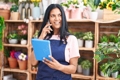 Middle age hispanic woman florist talking on smartphone using touchpad at florist