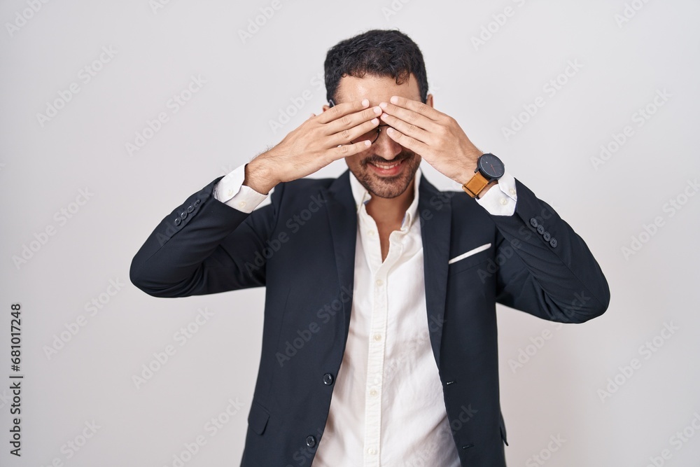 Handsome business hispanic man standing over white background covering eyes with hands smiling cheerful and funny. blind concept. Handsome business hispanic man standing over white background covering eyes with hands smiling cheerful and funny. blind concept.