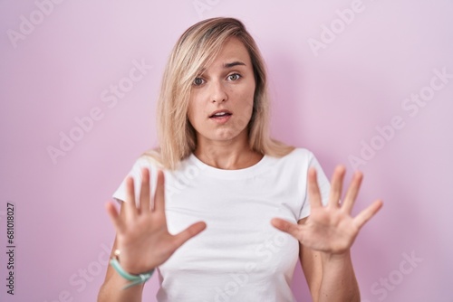 Young blonde woman standing over pink background afraid and terrified with fear expression stop gesture with hands, shouting in shock. panic concept. © Krakenimages.com