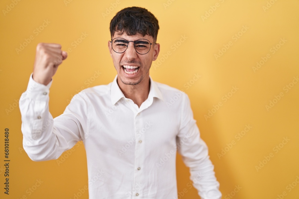 Young hispanic man standing over yellow background angry and mad raising fist frustrated and furious while shouting with anger. rage and aggressive concept. Young hispanic man standing over yellow background angry and mad raising fist frustrated and furious while shouting with anger. rage and aggressive concept.