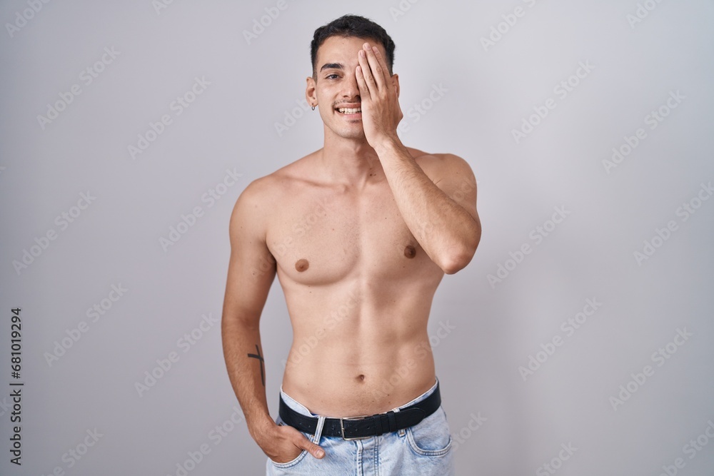 Fototapeta premium Handsome hispanic man standing shirtless covering one eye with hand, confident smile on face and surprise emotion.