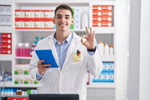 Handsome hispanic man working at pharmacy drugstore with tablet doing ok sign with fingers, smiling friendly gesturing excellent symbol