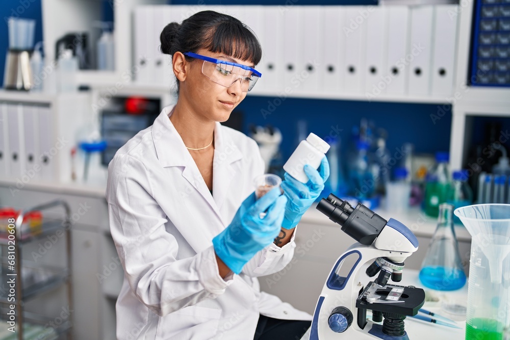 Young beautiful hispanic woman scientist smiling confident holding pills bottle at laboratory