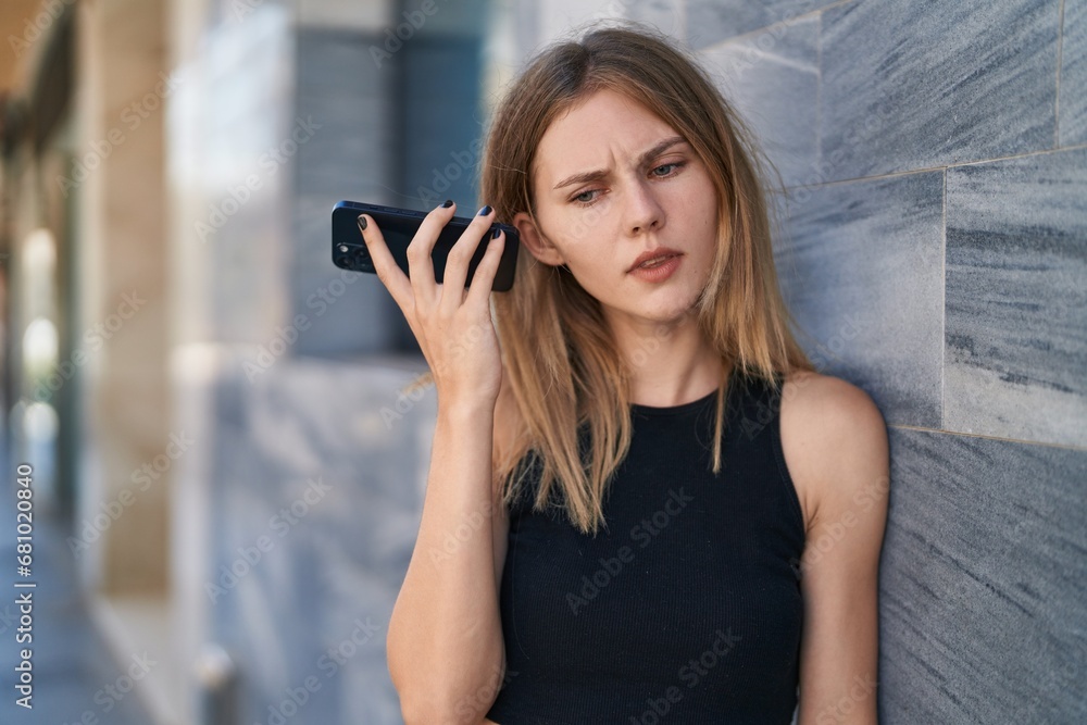 Fototapeta premium Young blonde woman listening to voice message with serious face at street