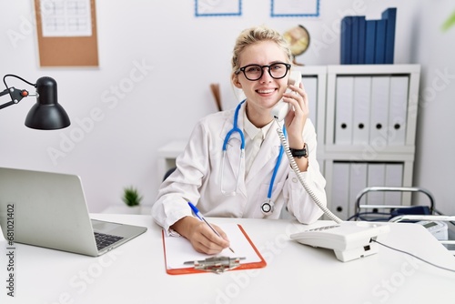 Young blonde woman doctor talking on telephone writing on document at clinic