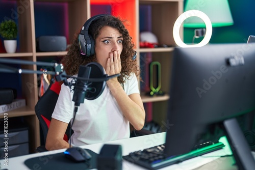 Hispanic woman with curly hair playing video games covering mouth with hand, shocked and afraid for mistake. surprised expression
