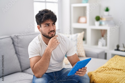 Bilde på lerret Hispanic man with beard using touchpad sitting on the sofa thinking worried abou