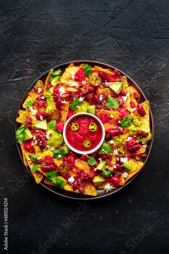 Loaded nachos. Mexican nacho chips with beef, overhead flat lay shot with guacamole sauce, cheese salsa, and chili peppers, on a black stone background