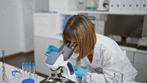 Relaxed yet focused, a young, beautiful hispanic woman scientist working diligently indoors at the lab, using a microscope. she's deeply engrossed in medical research amidst test tubes on the table.