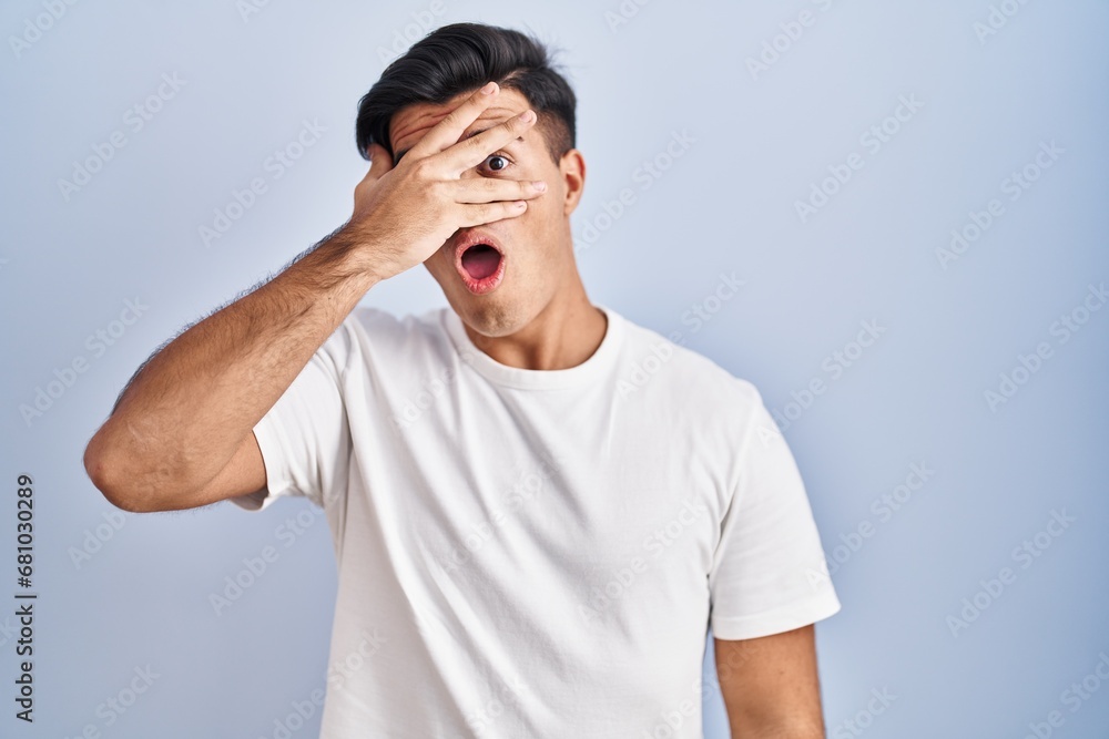 Hispanic man standing over blue background peeking in shock covering face and eyes with hand, looking through fingers with embarrassed expression. Hispanic man standing over blue background peeking in shock covering face and eyes with hand, looking through fingers with embarrassed expression.