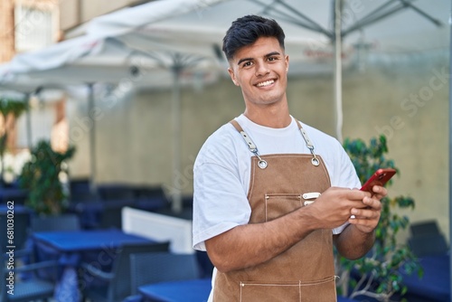 Young hispanic man waiter smiling confident using smartphone at coffee shop terrace