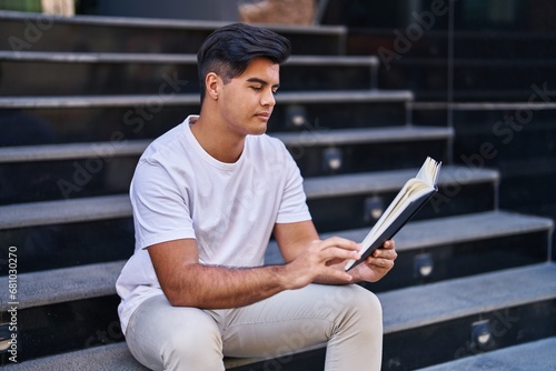 Young hispanic man reading book sitting on stairs at street