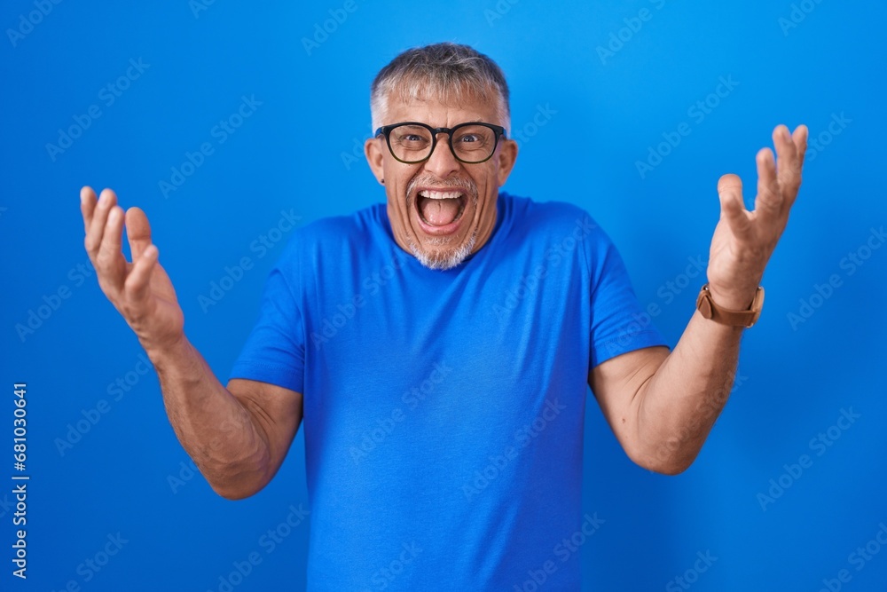 Hispanic man with grey hair standing over blue background celebrating mad and crazy for success with arms raised and closed eyes screaming excited. winner concept Hispanic man with grey hair standing over blue background celebrating mad and crazy for success with arms raised and closed eyes screaming excited. winner concept