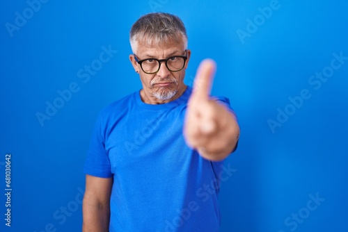 Hispanic man with grey hair standing over blue background pointing with finger up and angry expression, showing no gesture
