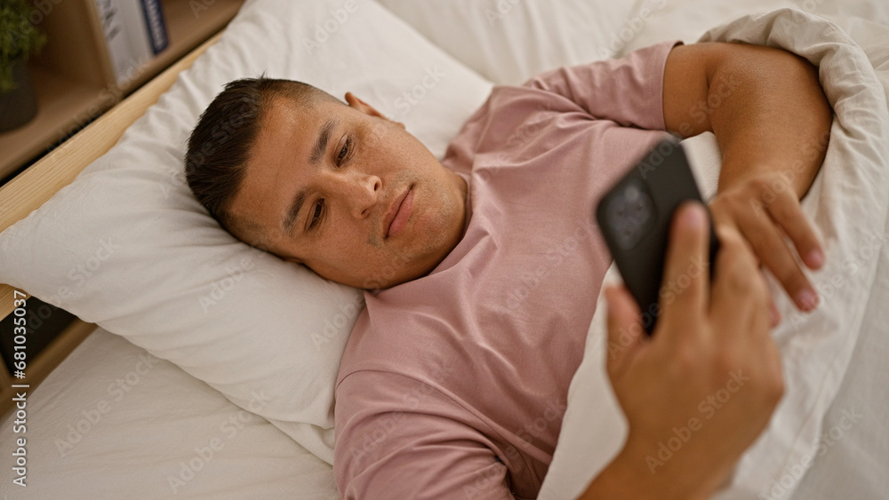 Young latin man using smartphone lying on bed at bedroom Young latin man using smartphone lying on bed at bedroom