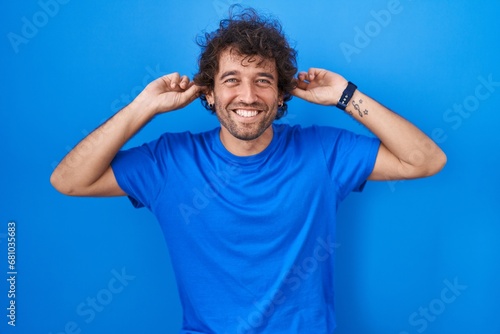 Hispanic young man standing over blue background smiling pulling ears with fingers, funny gesture. audition problem