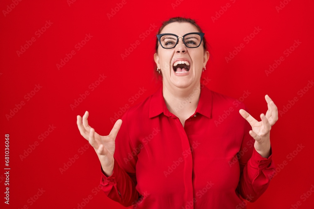 Naklejka premium Young hispanic woman with red hair standing over red background crazy and mad shouting and yelling with aggressive expression and arms raised. frustration concept.