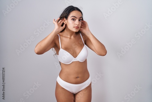 Young hispanic woman wearing white lingerie trying to hear both hands on ear gesture, curious for gossip. hearing problem, deaf © Krakenimages.com