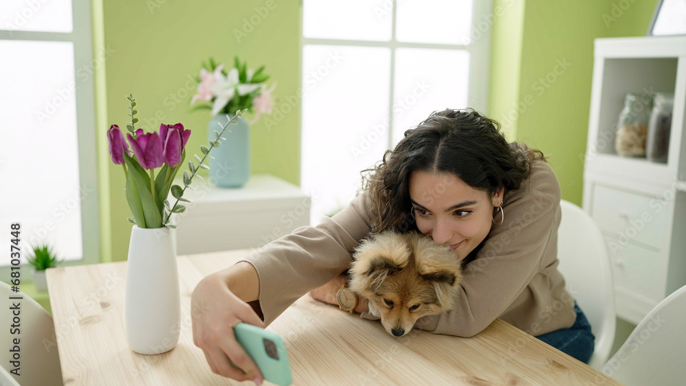 Young hispanic woman with dog sitting on table make selfie by smartphone at dinning room