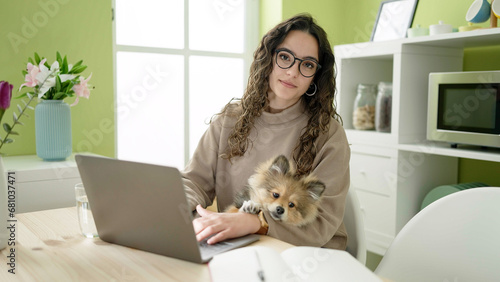 Young hispanic woman with dog sitting on table using laptop at dinning room