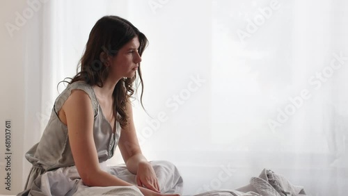 A young woman in a gray nightgown is sitting on the bed and thinking about something. A woman who woke up early in the morning sits on the bed and mentally prepares for a hard working day.