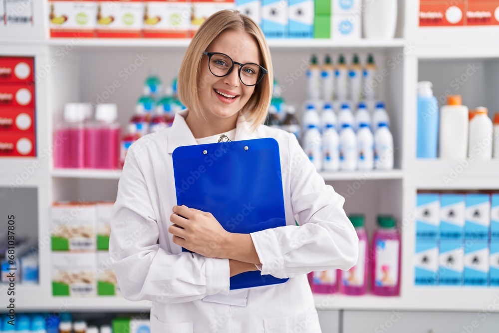 Young blonde woman pharmacist smiling confident holding clipboard at pharmacy