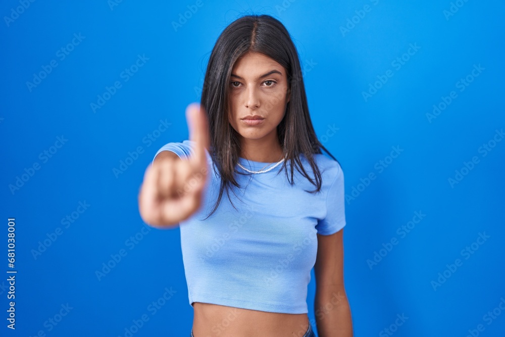 Brunette young woman standing over blue background pointing with finger up and angry expression, showing no gesture Brunette young woman standing over blue background pointing with finger up and angry expression, showing no gesture