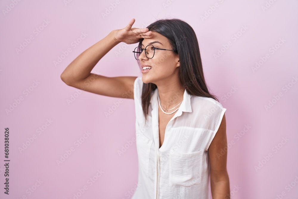 Brunette young woman standing over pink background wearing glasses very happy and smiling looking far away with hand over head. searching concept. Brunette young woman standing over pink background wearing glasses very happy and smiling looking far away with hand over head. searching concept.