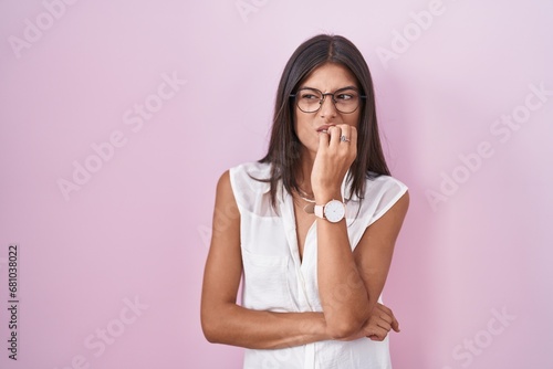 Brunette young woman standing over pink background wearing glasses looking stressed and nervous with hands on mouth biting nails. anxiety problem. photo