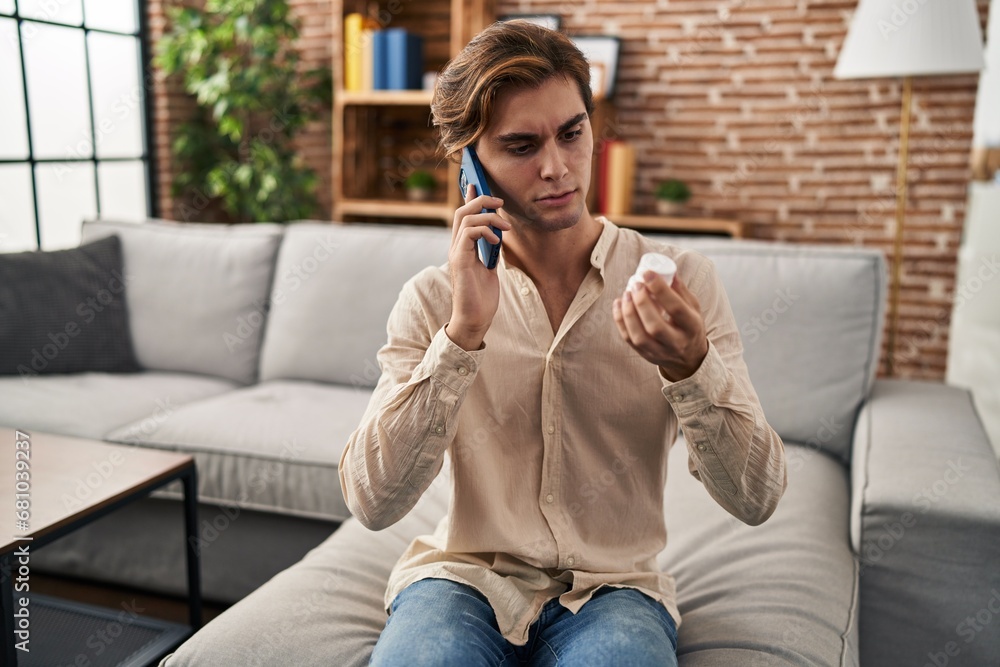 Fototapeta premium Young caucasian man talking on smartphone holding pills bottle at home
