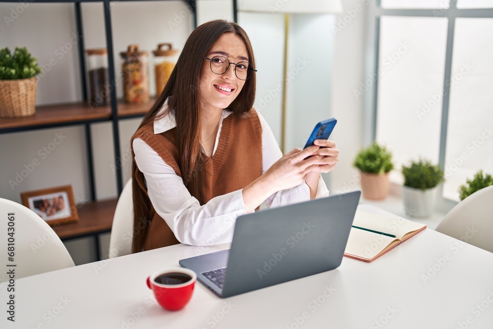 Young beautiful hispanic woman using smartphone and laptop sitting on table at home Young beautiful hispanic woman using smartphone and laptop sitting on table at home