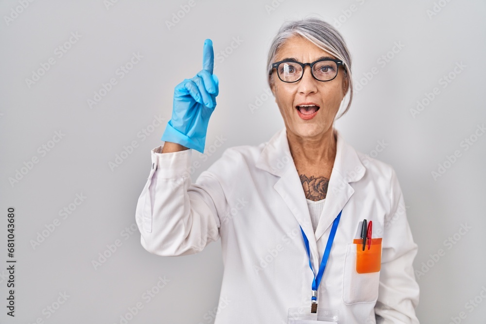 Middle age woman with grey hair wearing scientist robe pointing finger up with successful idea. exited and happy. number one. Middle age woman with grey hair wearing scientist robe pointing finger up with successful idea. exited and happy. number one.