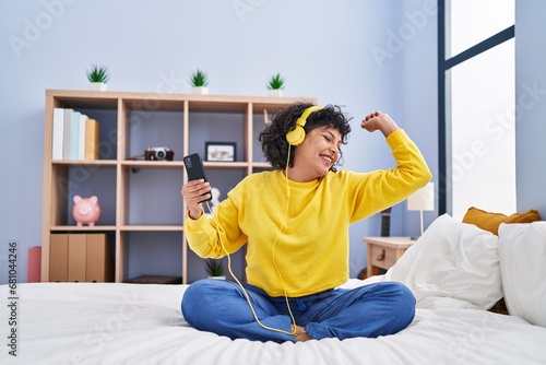 Young beautiful hispanic woman dancing and listening to music sitting on bed at bedroom